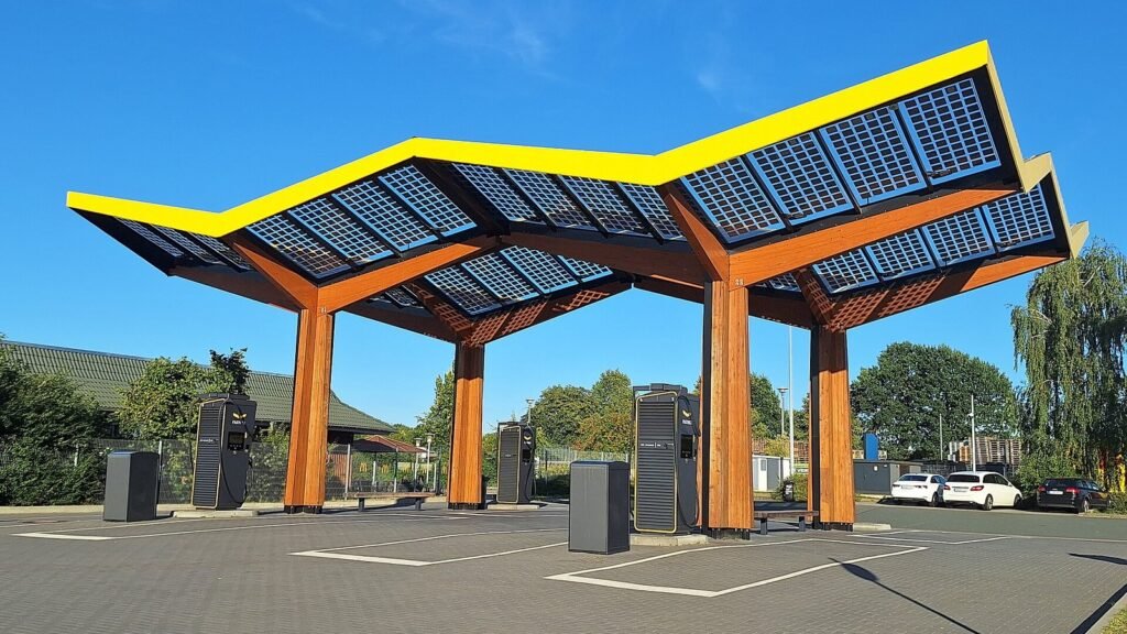 Station de recharge rapide pour voitures électriques sous un auvent en bois couvert de panneaux solaires, avec plusieurs bornes sur une aire de stationnement, ciel bleu