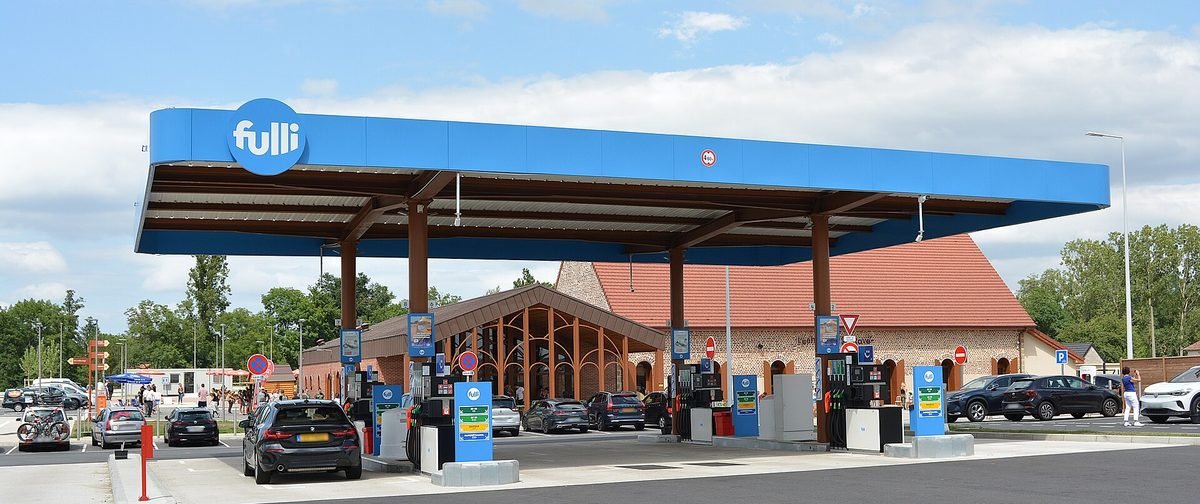 Station-service Fulli de l’aire du Bourbonnais sur l’A79, avec son grand auvent bleu, ses pompes et quelques voitures, sous un ciel nuageux