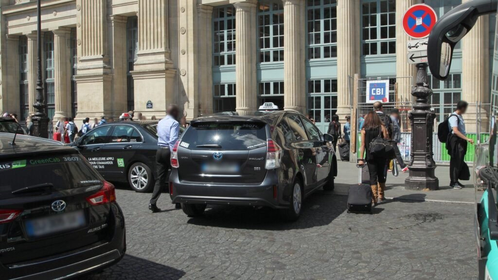 Taxis parisiens garés devant la façade à colonnes de la gare du Nord à Paris, des voyageurs avec valises sur le trottoir, par une journée ensoleillée
