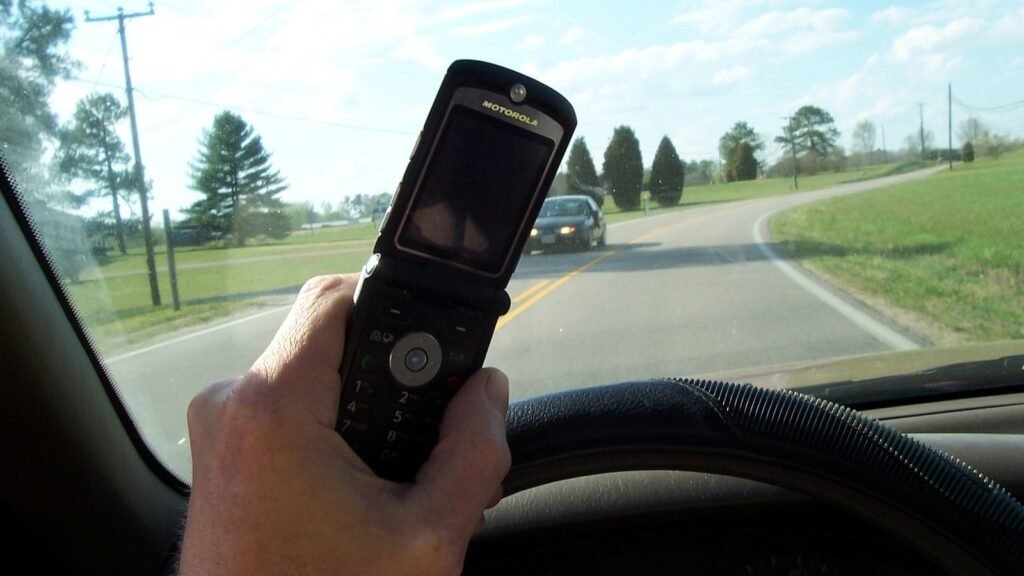Main de conducteur tenant un téléphone à clapet devant le volant, avec une route de campagne et une voiture au loin