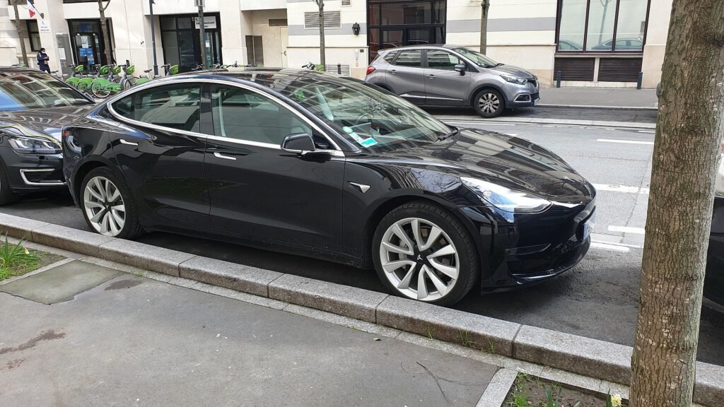 Tesla Model 3 noire stationnée le long d’un trottoir dans une rue de la région parisienne, avec des vélos en libre-service et des immeubles en arrière-plan