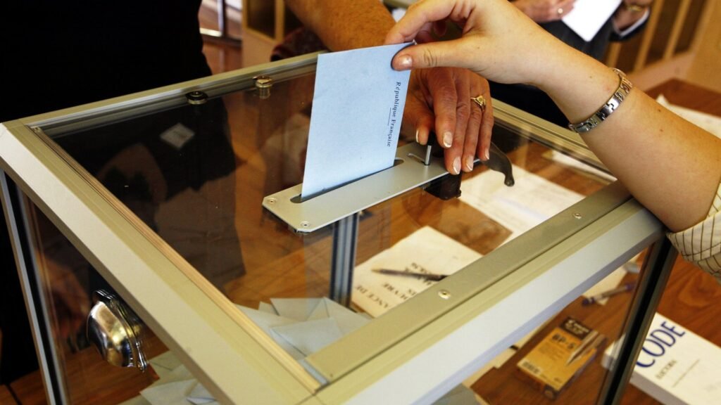 Une main glisse une enveloppe de vote dans une urne transparente posée sur une table de bureau de vote