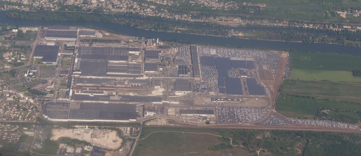 Vue aérienne de l’usine Renault de Flins-sur-Seine au bord de la Seine, avec ses grands bâtiments et des parkings remplis de voitures neuves