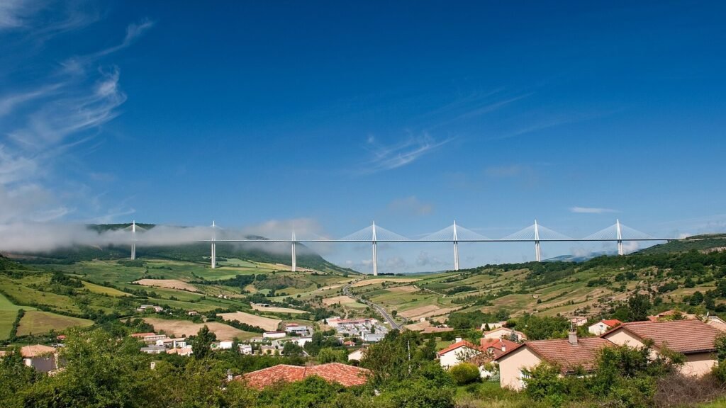 Le viaduc de Millau et ses piles blanches à haubans tendu au-dessus de collines vertes, sous un ciel bleu, avec les toits de tuiles du village de Creissels au premier plan