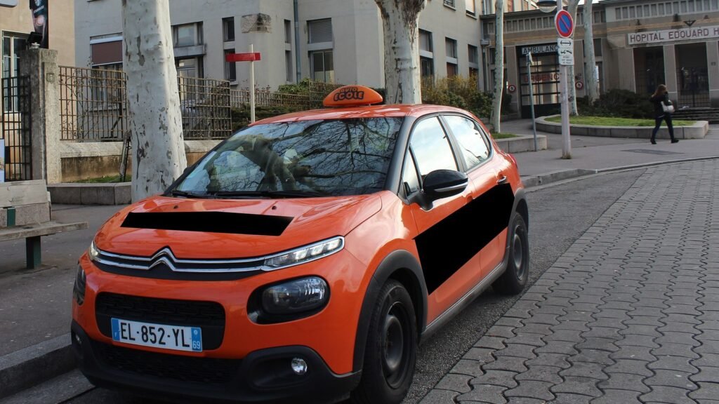 Une Citroën C3 orange d’auto-école, avec le panneau « École » sur le toit, garée le long d’un trottoir pavé devant un bâtiment