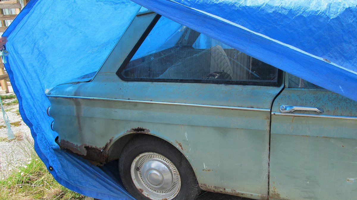 Vieille voiture bleu-vert à la carrosserie rouillée, à moitié recouverte d’une bâche plastique bleue