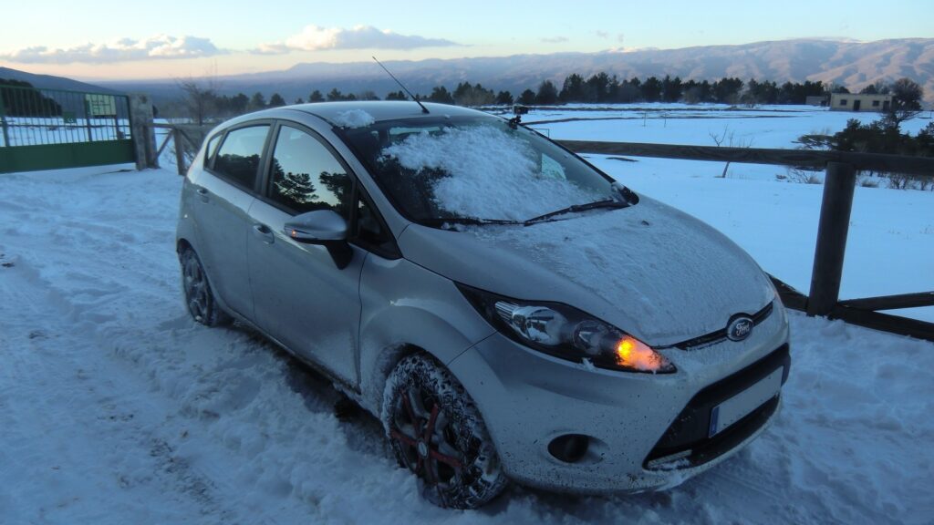 Ford Fiesta grise couverte de neige, avec des chaînes à neige montées sur la roue avant, sur un chemin enneigé devant un paysage de montagne au crépuscule