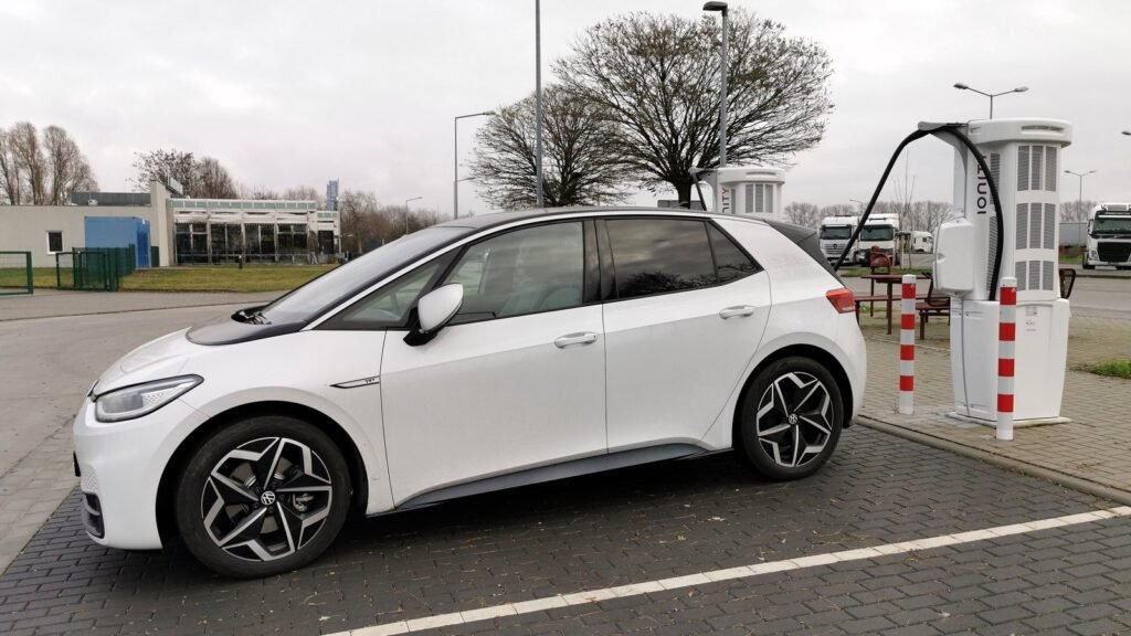 Volkswagen ID.3 blanche garée à une borne de recharge rapide Ionity sur une aire d’autoroute allemande, sous un ciel gris d’hiver, arbres sans feuilles