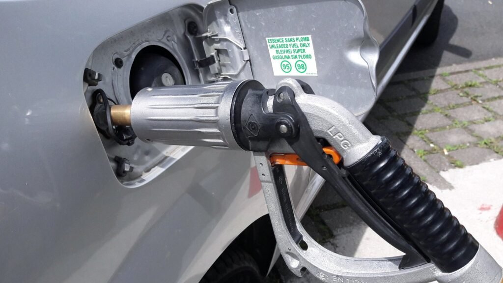 Pistolet de GPL raccordé à un adaptateur vissé dans la trappe à carburant d’une voiture grise, à côté de l’étiquette « essence sans plomb »