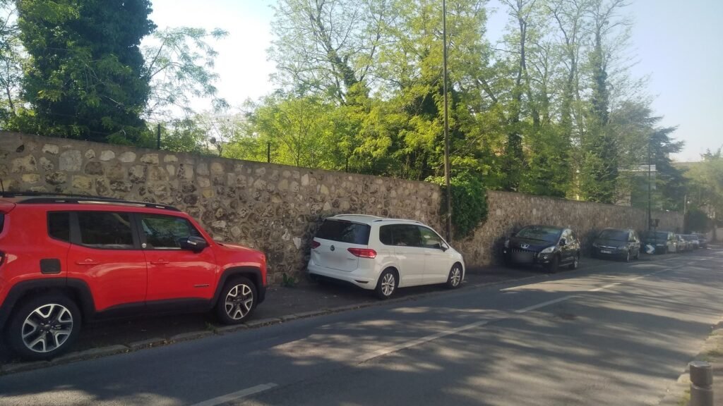 Rue bordée d’un mur de pierre où plusieurs voitures, dont un SUV rouge et un monospace blanc, sont garées avec deux roues sur le trottoir, au soleil